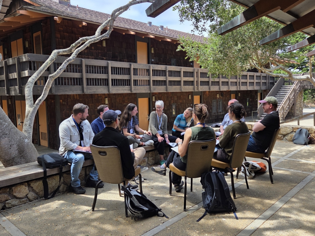 A group of people seated in a circle outdoors under an oak tree, in discussion during a conference breakout session at a rustic lodge.