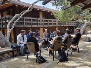 A group of people seated in a circle outdoors under an oak tree, in discussion during a conference breakout session at a rustic lodge.