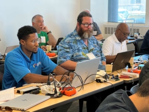 Three men concentrating on their laptops during a workshop, with various notes, devices, and tools on the table in front of them.